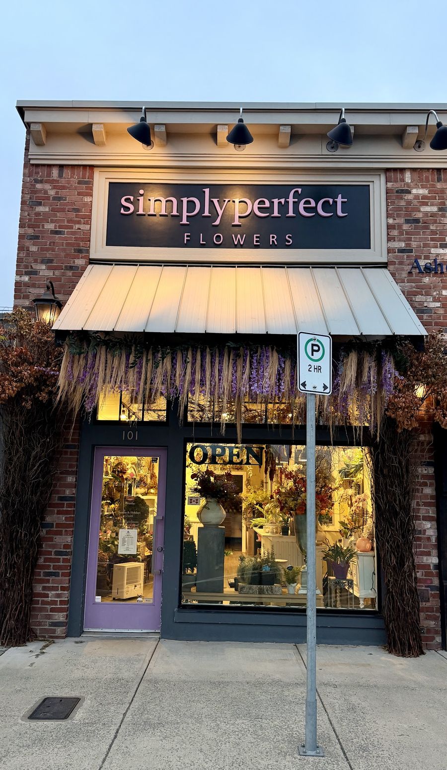 Simply Perfect Flowers storefront at dusk, windows lit, dried flowers hanging over the door